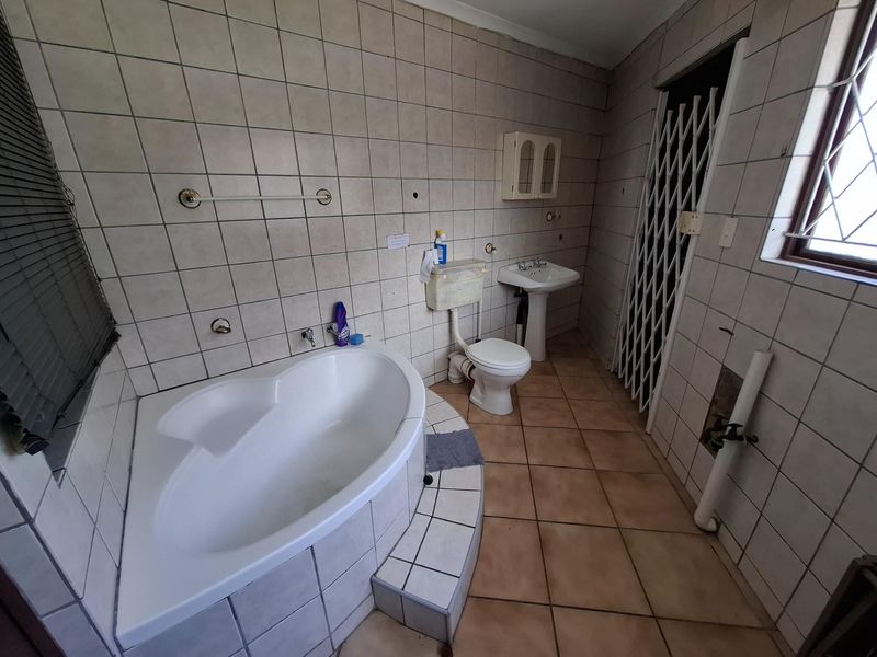 A dated bathroom featuring a corner bathtub, pedestal basin, and toilet, with full-height wall tiling and ceramic floor tiles.