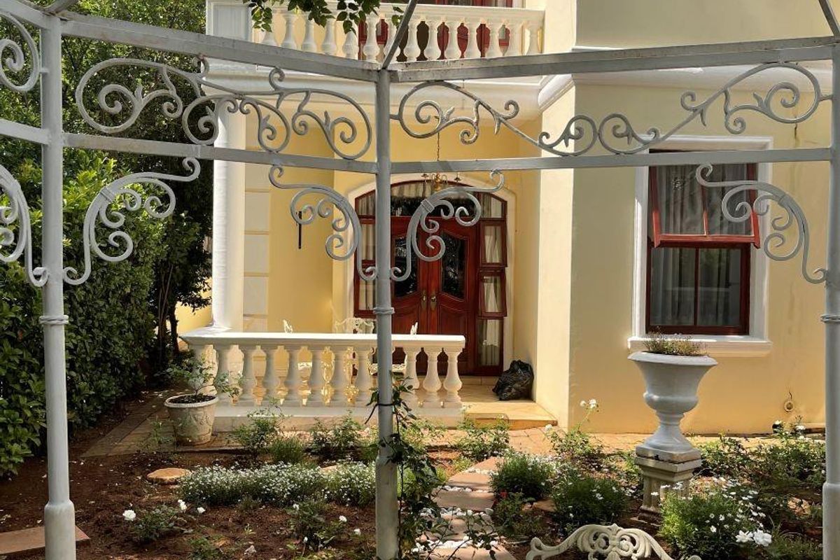 A charming rear garden view featuring a paved pathway, landscaped flower beds, and a classic two-story building facade with a balcony and decorative balustrades.