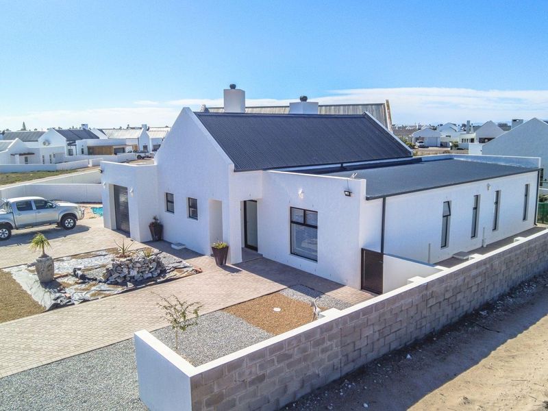 Aerial view of a modern white house with a dark corrugated metal roof, featuring a paved driveway and perimeter boundary wall in a coastal estate.