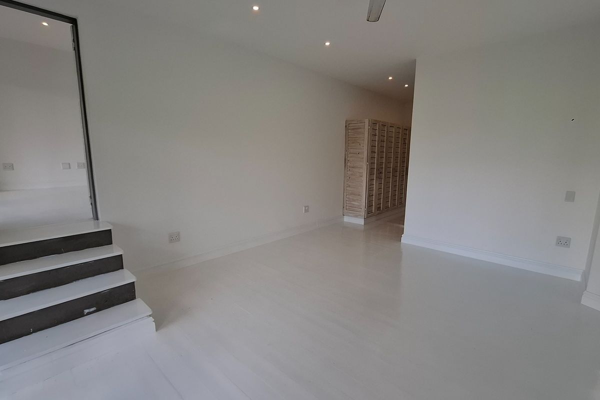 A bright, open-plan living area featuring white painted flooring, recessed lighting, and a small internal staircase leading to an adjacent space.
