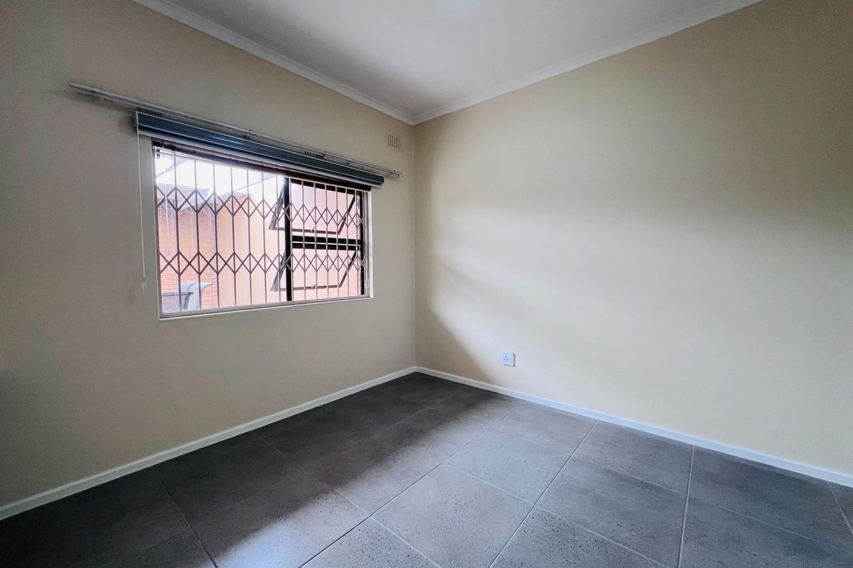 A clean, empty bedroom featuring neutral wall paint, large grey floor tiles, and a window fitted with a security gate and roller blind.