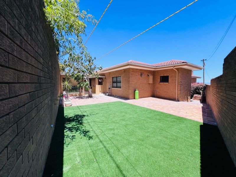 Rear exterior view of the apartment featuring a brick facade, tiled roof, paved patio area, and a well-maintained artificial lawn enclosed by boundary walls.
