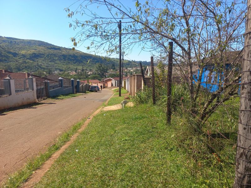 A street view showing a residential road with surrounding properties, wire fencing, and a scenic mountain backdrop.