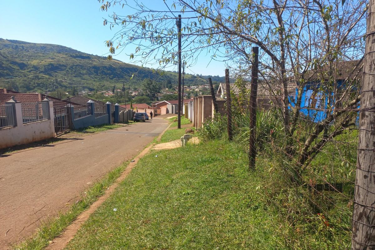 A street view showing a residential road with surrounding properties, wire fencing, and a scenic mountain backdrop.