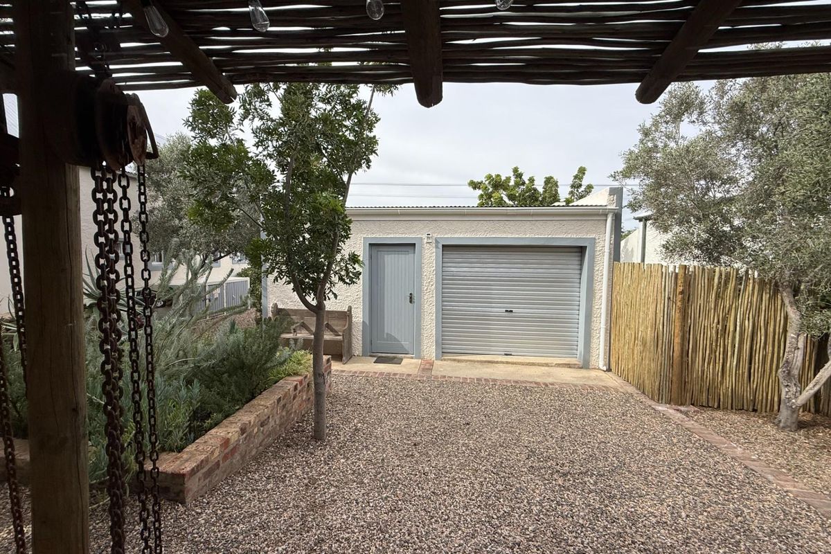 A view of a detached garage with a roller door and side entrance, set within a landscaped garden area featuring a timber pergola.