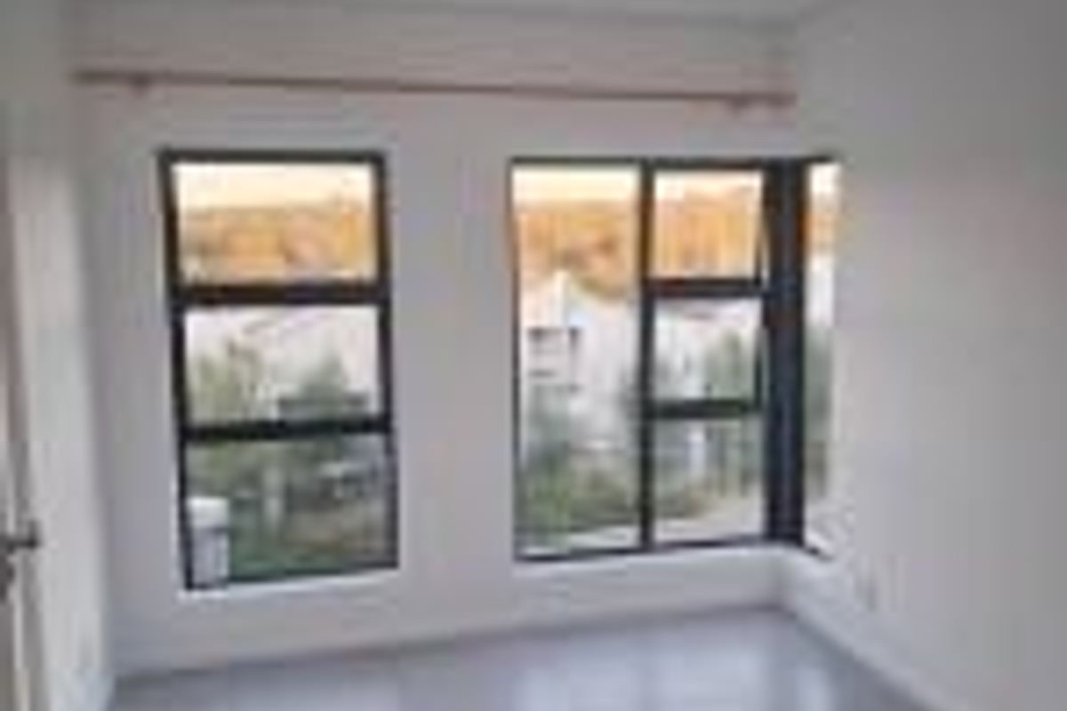 A bright, empty bedroom featuring large windows with dark frames, neutral tiled flooring, and recessed ceiling lighting.