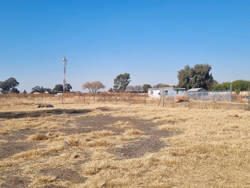 A wide-angle view of a large, undeveloped farm plot featuring dry grass, perimeter fencing, and distant outbuildings under a clear blue sky.