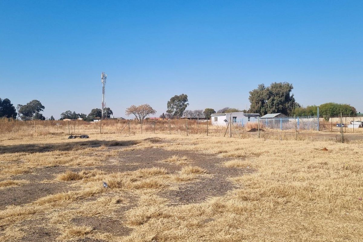 A wide-angle view of a large, undeveloped farm plot featuring dry grass, perimeter fencing, and distant outbuildings under a clear blue sky.