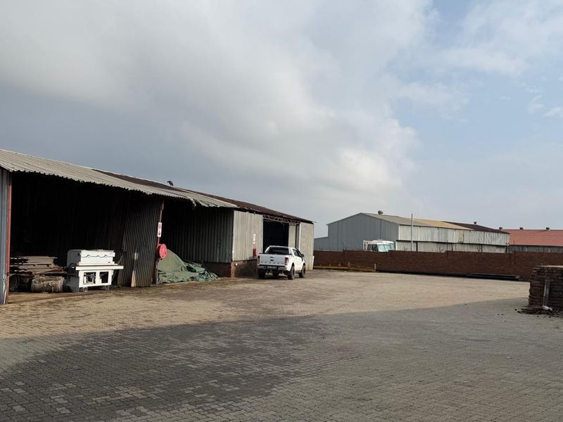 Industrial yard featuring large corrugated metal storage sheds, paved driveway area, and perimeter brick walling.