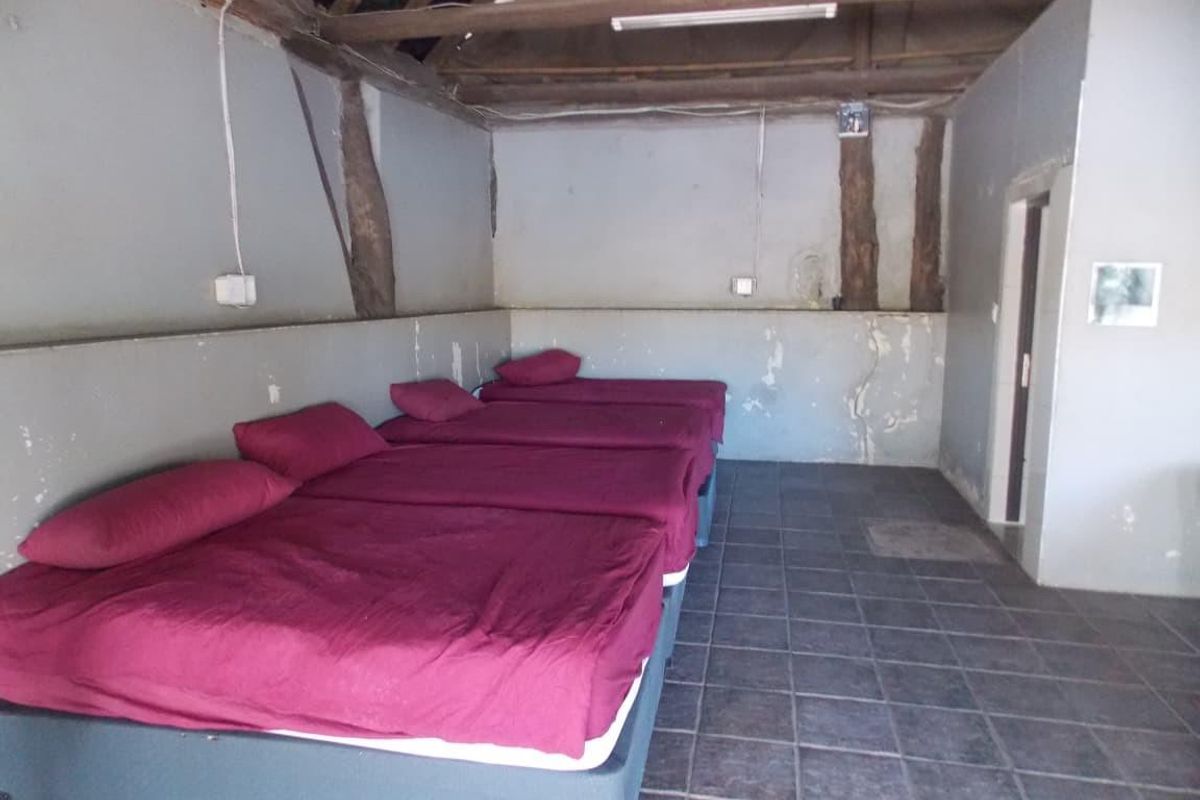 A rustic bedroom featuring tiled flooring, exposed timber roof beams, and multiple bed placements within a simple, utilitarian space.