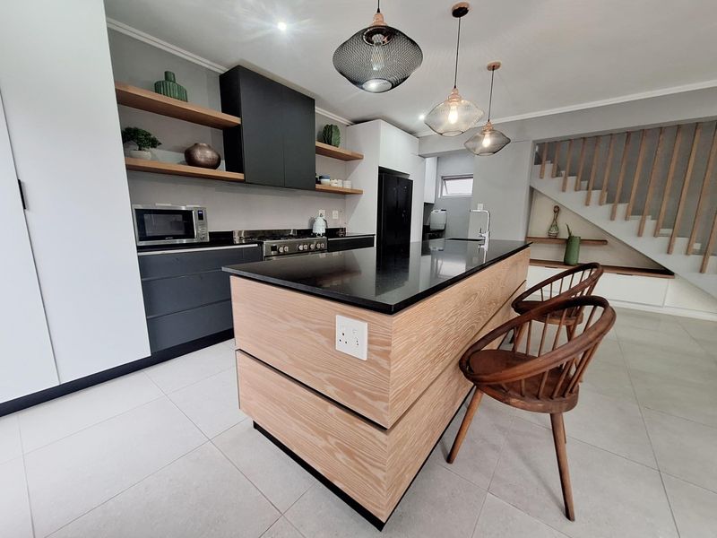 Modern kitchen featuring a central island with stone countertops, built-in appliances, floating shelving, and an adjacent staircase with a timber balustrade.