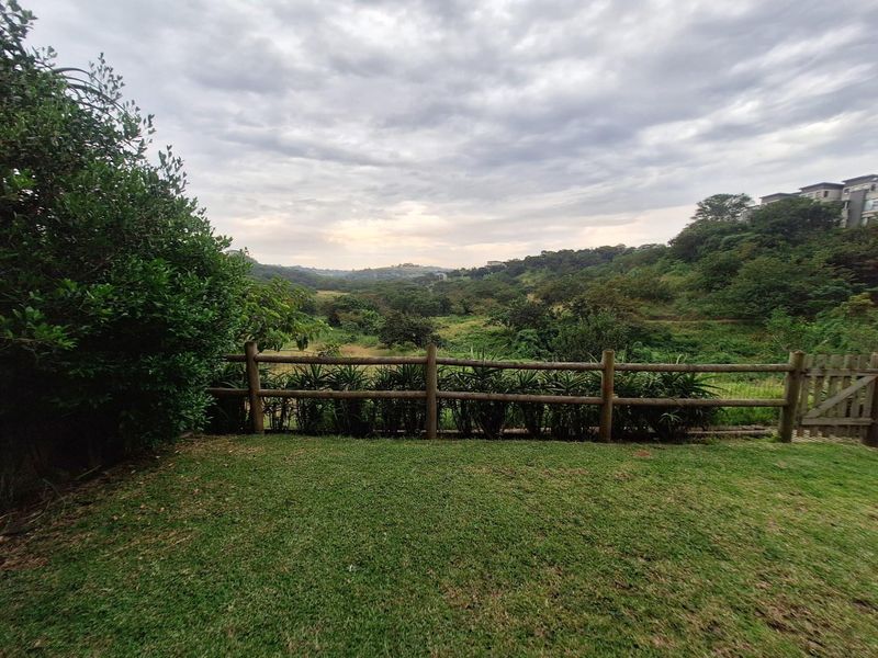 A scenic view overlooking a lush green valley and natural landscape, framed by a timber post and rail fence and well-maintained lawn.