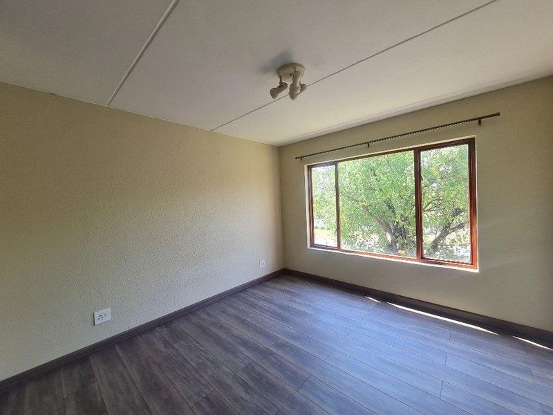 A bright, empty bedroom featuring wood-look laminate flooring, neutral textured walls, and a large window with a view of mature trees.