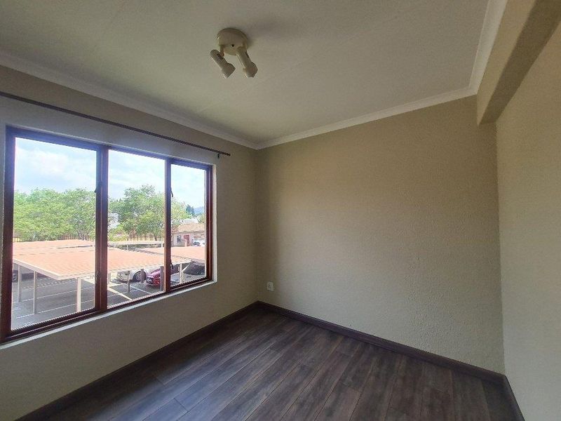 A bright, empty bedroom featuring dark wood-look laminate flooring, neutral walls, and large windows overlooking the complex parking area.