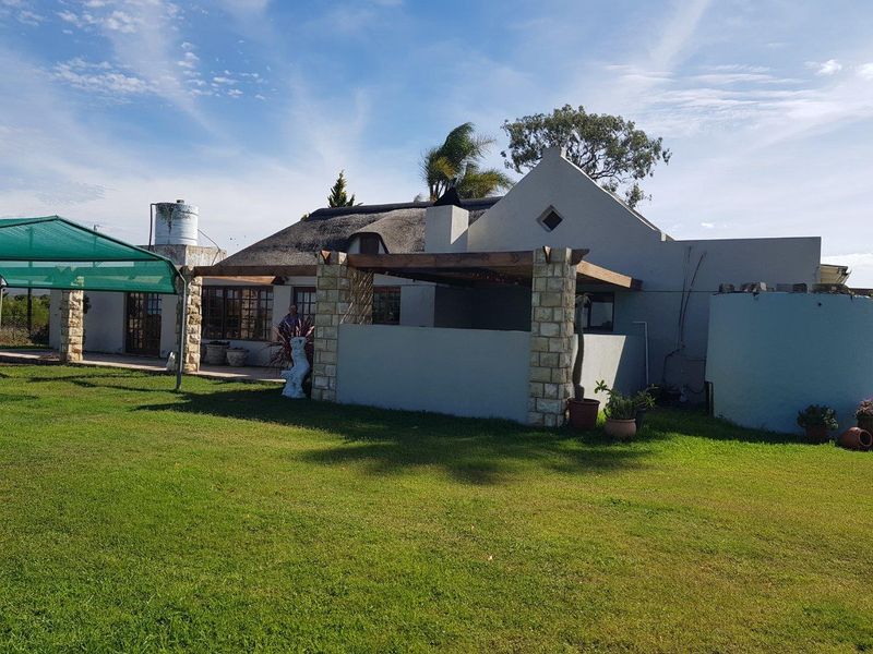 Rear view of a farm-style residence featuring a thatched roof, stone-clad pillars, and a covered patio area overlooking a spacious lawn.