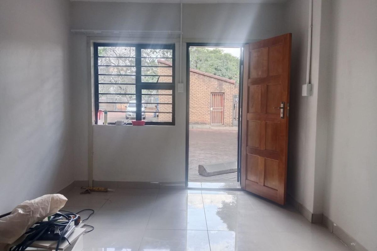 A bright, empty room featuring tiled flooring, a wooden entrance door, and a window with security bars, providing direct access to the exterior.