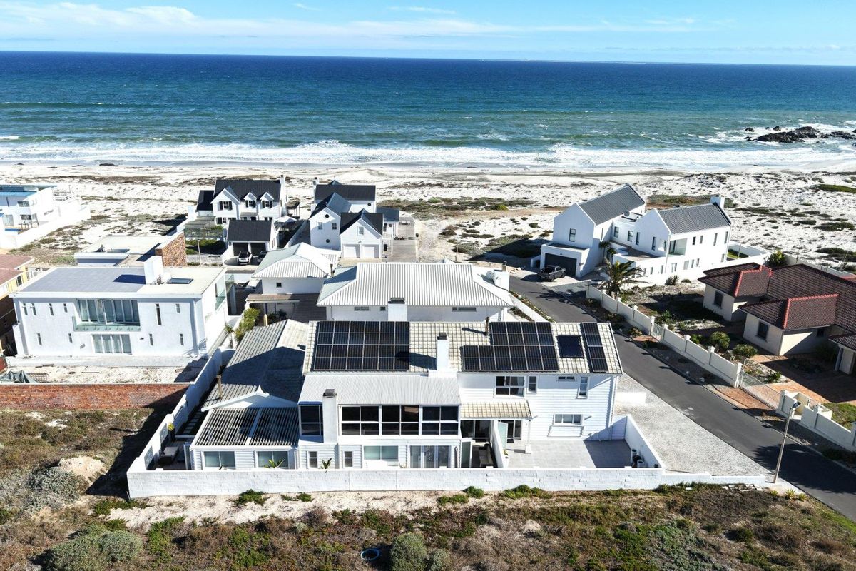 Aerial view of a coastal residential property featuring a white exterior, metal roof, and extensive solar panel installation, situated near the ocean.