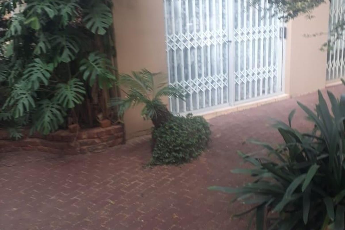 A paved rear patio area featuring a covered section and security gates leading into the home, surrounded by established garden beds.