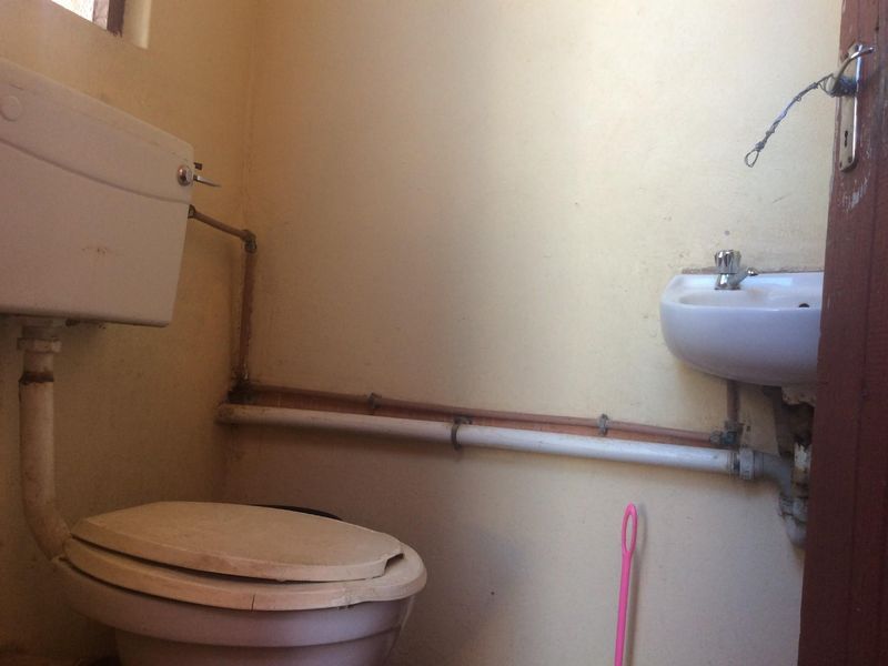 A small, basic bathroom featuring a toilet and wall-mounted basin with exposed plumbing, requiring cosmetic updates and maintenance to improve its overall condition.