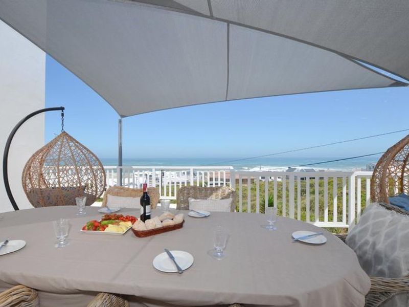 A covered outdoor patio area featuring a shade sail, white balustrade railing, and expansive ocean views, perfect for alfresco dining.