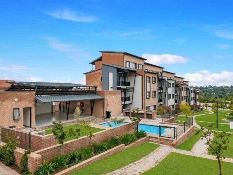 Exterior view of a modern apartment complex featuring a communal swimming pool, landscaped gardens, and a covered outdoor entertainment area with brick finishes.