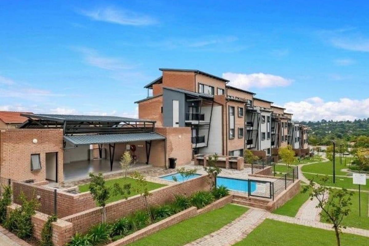 Exterior view of a modern apartment complex featuring a communal swimming pool, landscaped gardens, and a covered outdoor entertainment area with brick finishes.