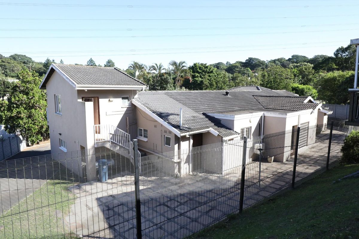 An elevated view of the rear exterior of the house, featuring a multi-level roof structure, paved courtyard area, and perimeter security fencing.