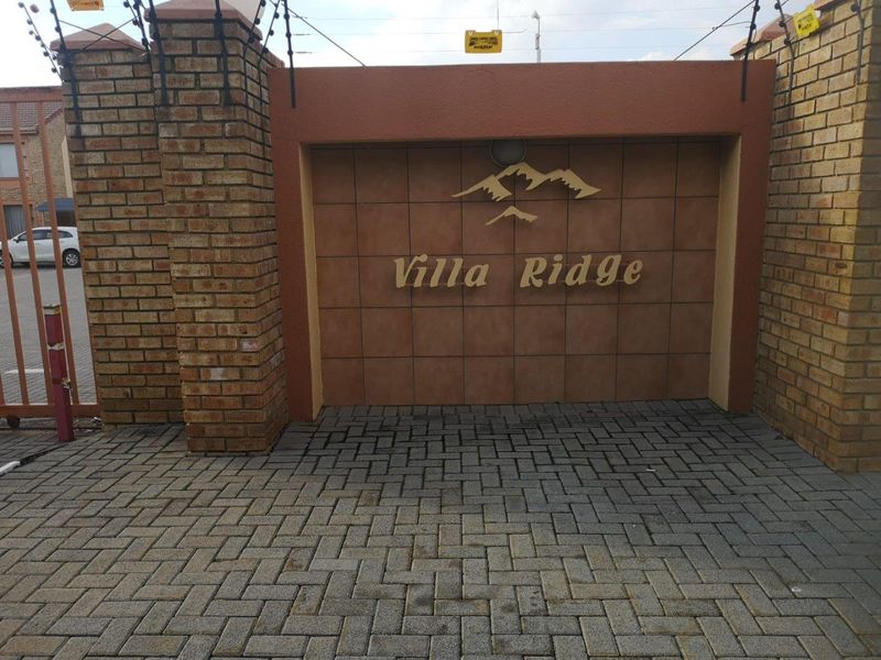 Entrance view of the Villa Ridge townhouse complex, featuring brick pillars, paved driveway, and security fencing.
