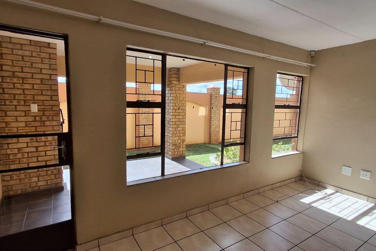 A bright, empty living area featuring large windows with security bars, tiled flooring, and direct access to an outdoor patio space.