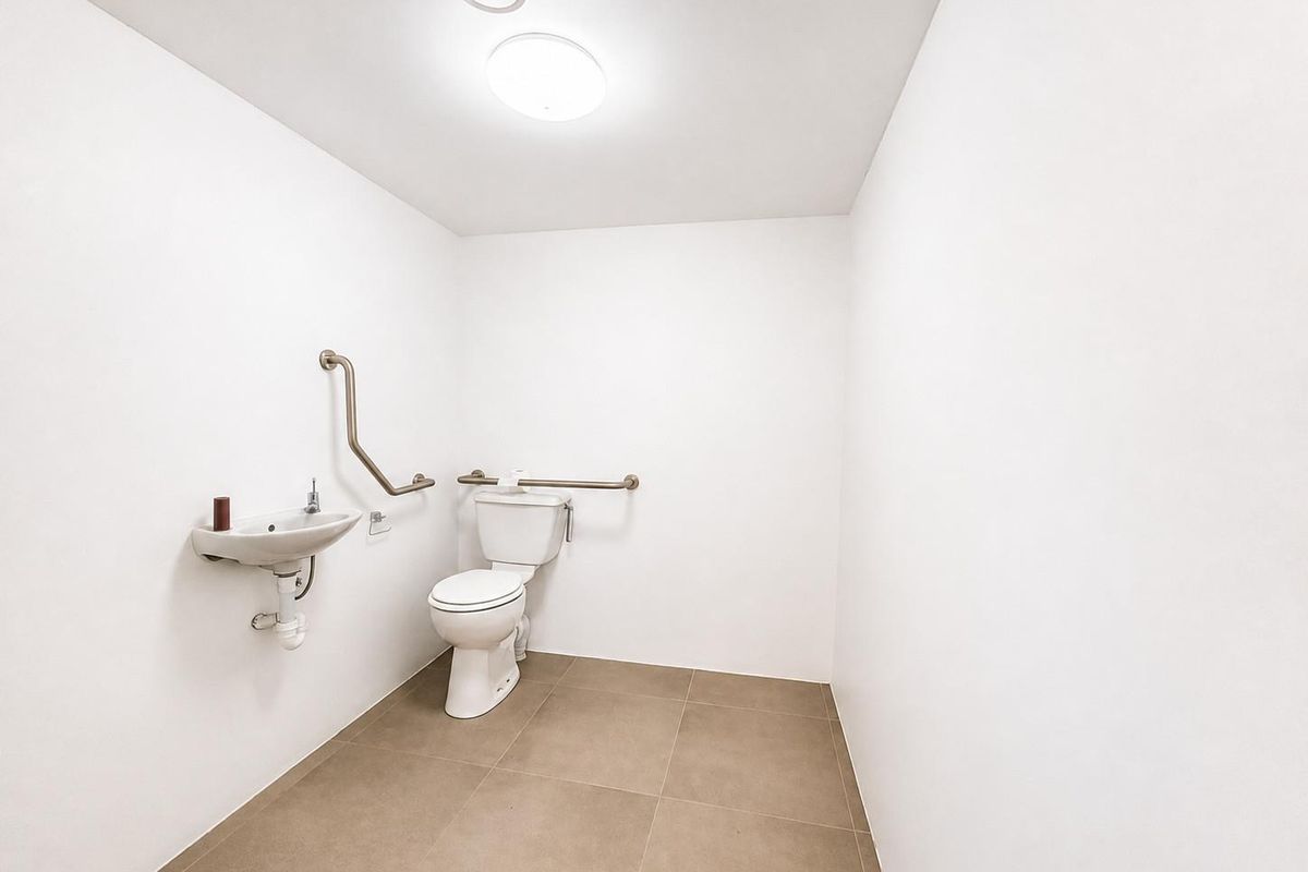 A clean, accessible bathroom featuring a wall-mounted basin, toilet, and stainless steel grab rails, finished with neutral floor tiling.