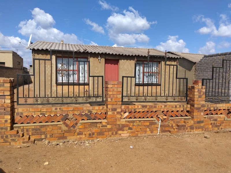 Front view of a single-story house featuring a brick boundary wall, metal security fencing, and a corrugated roof.