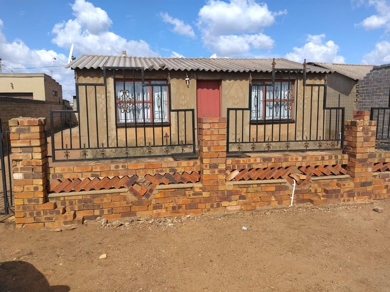 Front view of a single-story house featuring a brick boundary wall with metal security fencing and a simple facade with security bars on windows.