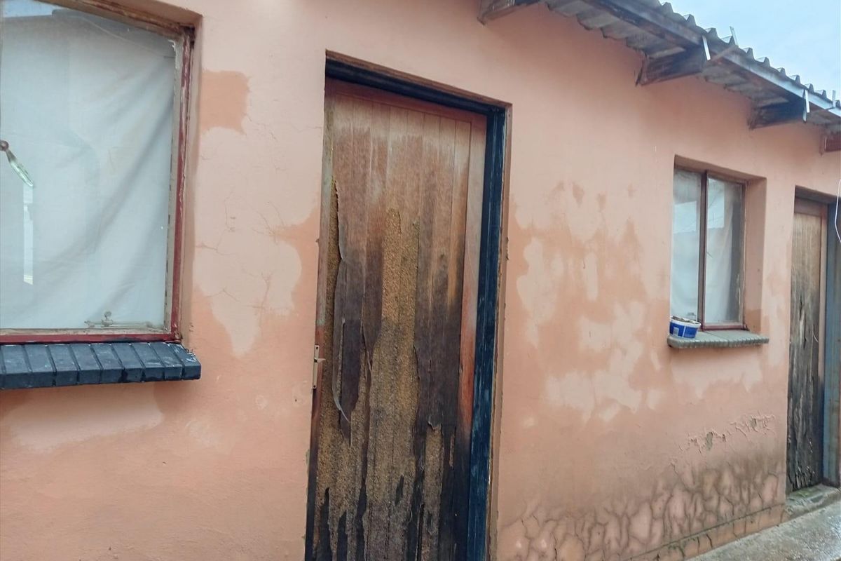 Exterior view of a residential building showing significant wear to the facade, including peeling paint, cracked plaster, and a damaged wooden door.