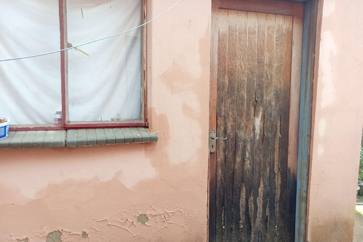 Exterior view of a building facade showing a weathered wooden door, peeling paint, and signs of structural deterioration on the wall and window sill.