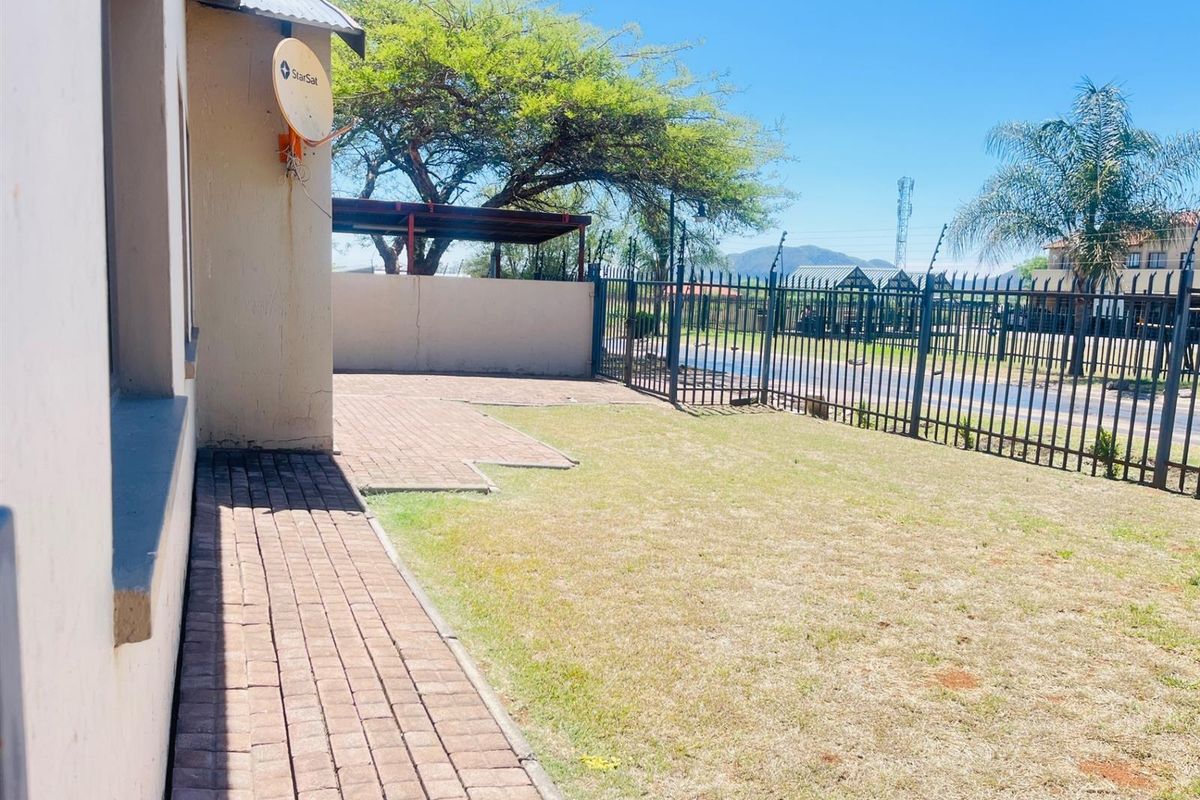A rear exterior view of the property featuring a paved walkway, a small lawn area, and secure perimeter fencing.