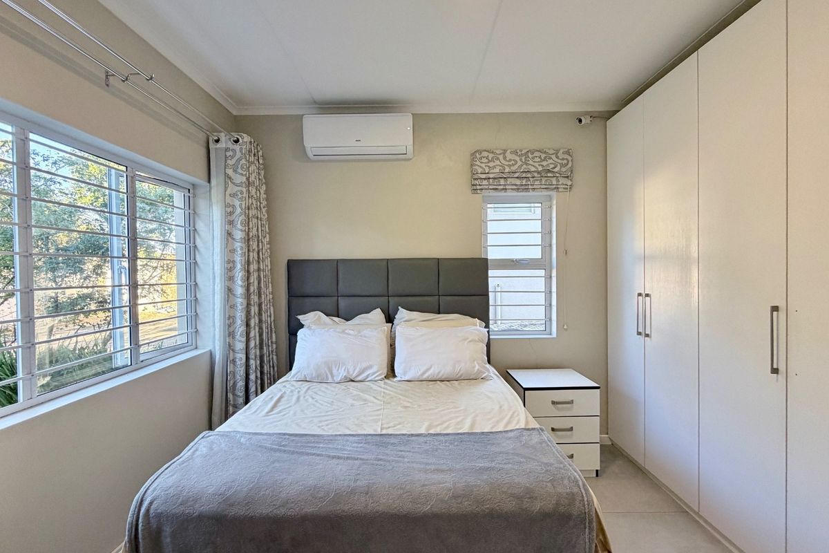 A well-maintained bedroom featuring built-in floor-to-ceiling wardrobes, a wall-mounted air conditioning unit, and large windows with security bars.