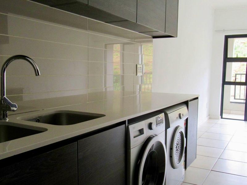Modern kitchen featuring dark cabinetry, stone countertops, stainless steel sink, and integrated laundry appliance space, leading to a balcony with natural light.