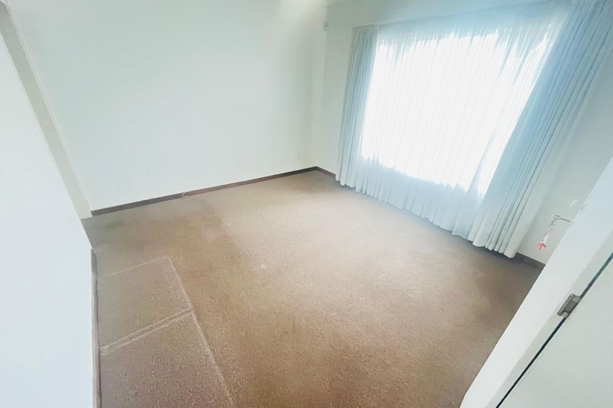A vacant bedroom featuring neutral walls and wall-to-wall carpeting, illuminated by a large window with fitted curtains.