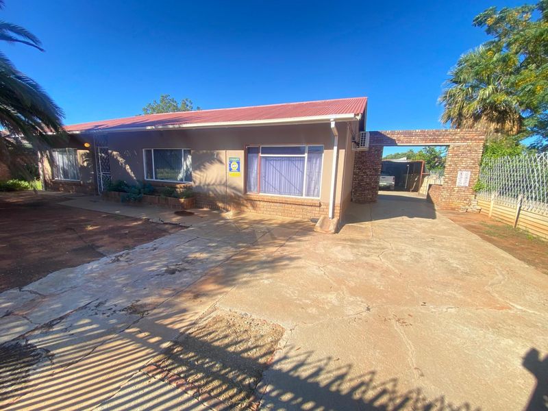 Front view of a single-story house featuring a red metal roof, brick facade, and a spacious concrete driveway leading to a side carport.