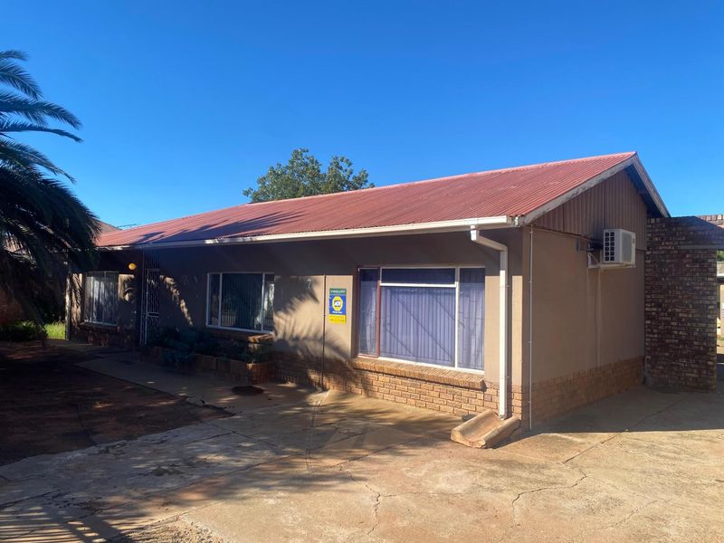 A single-story residential house featuring a corrugated metal roof, brick base detailing, and a paved driveway area.
