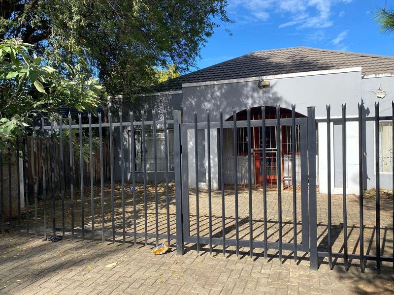 Front exterior view of a single-story house featuring a paved driveway, security fencing, and a gated entrance.