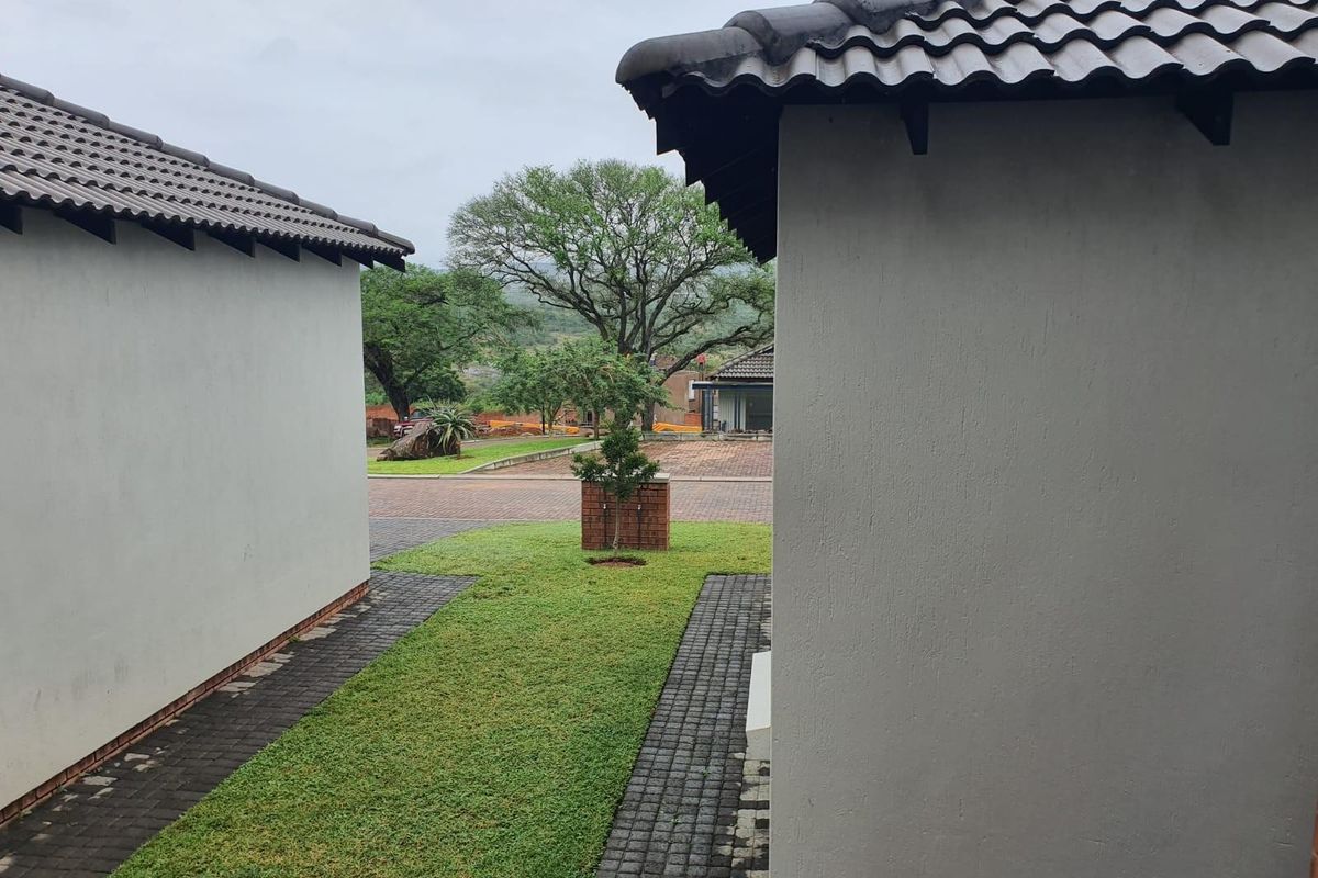 A view between two residential structures showing a paved walkway, manicured lawn, and a brick pillar feature in a landscaped garden setting.