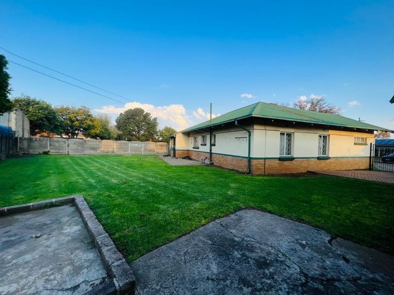 A spacious rear garden featuring a well-maintained lawn, concrete patio area, and a view of the house's rear elevation with a green metal roof.