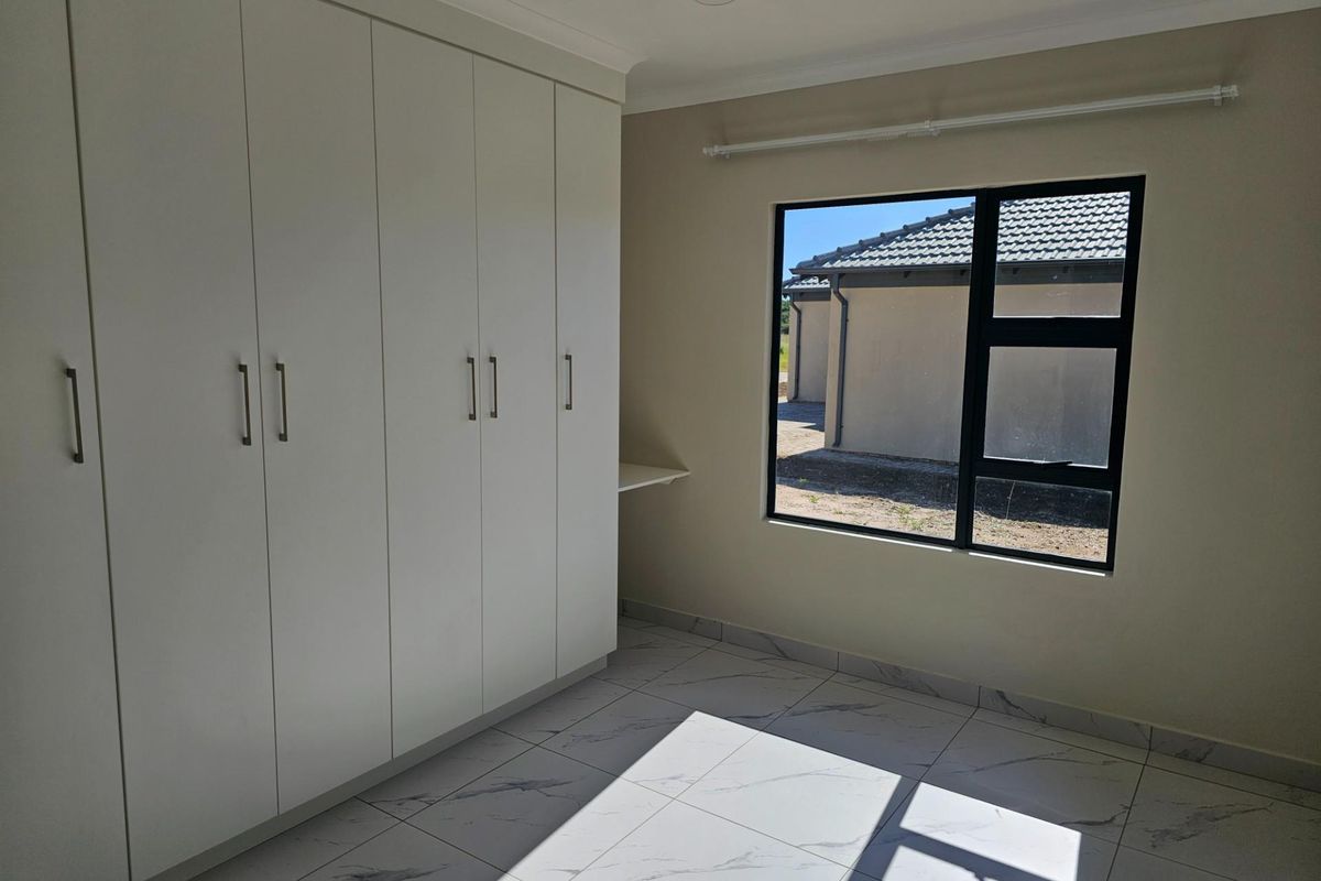 A bright bedroom featuring modern white built-in cupboards, large format marble-look floor tiles, and a window with a view of the neighboring property.