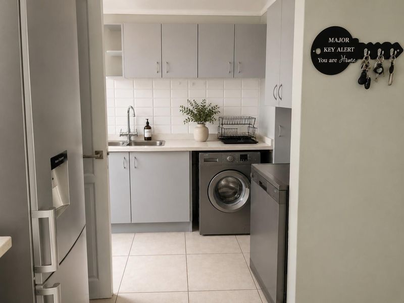 A functional laundry area featuring grey built-in cabinetry, a stainless steel sink, tiled backsplash, and tiled flooring with space for appliances.
