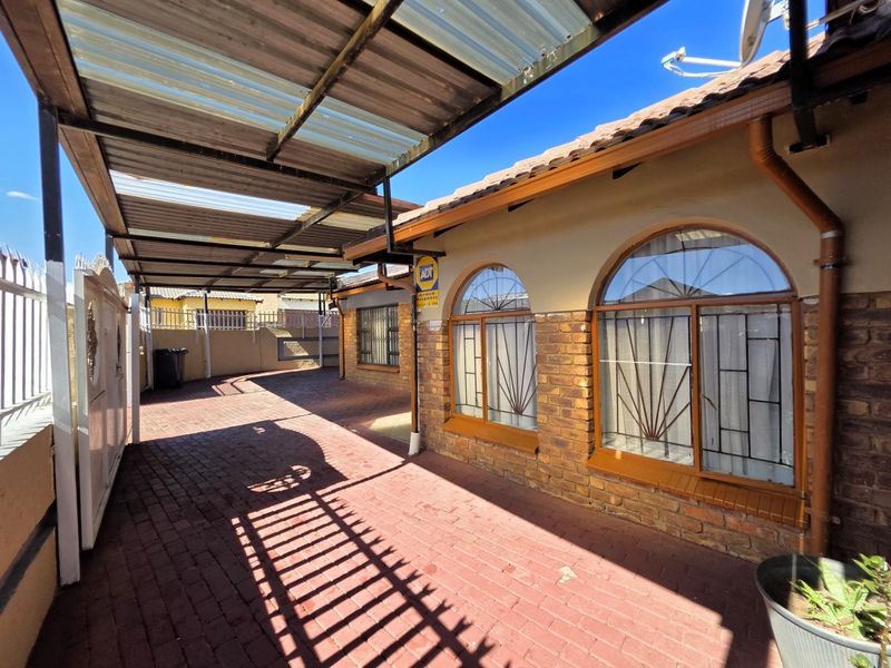 A paved front driveway featuring a covered carport, brick facade with arched windows, and secure perimeter walling.