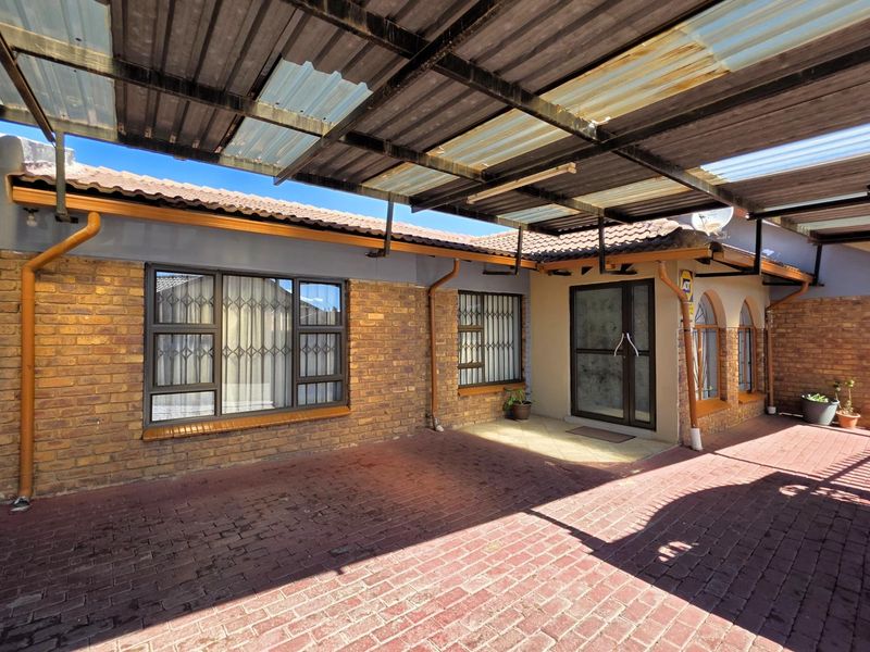 Front exterior view featuring a brick facade, paved driveway, and a covered carport area with security gates and aluminium window frames.