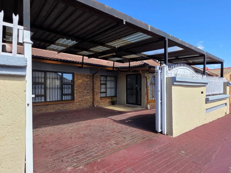 Front exterior view featuring a paved driveway, covered carport, and secure perimeter wall with metal security gates and window bars.