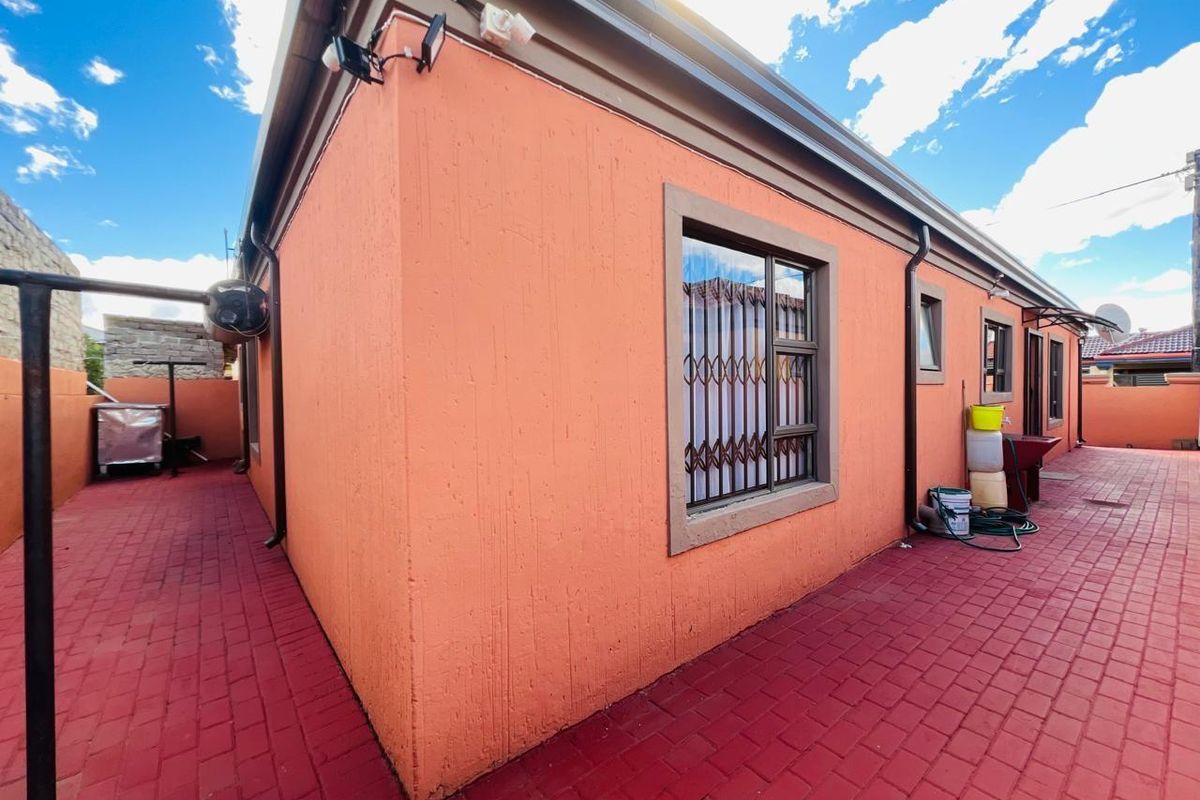 Side exterior view of the house featuring textured orange walls, security bars on windows, and red paved pathways.