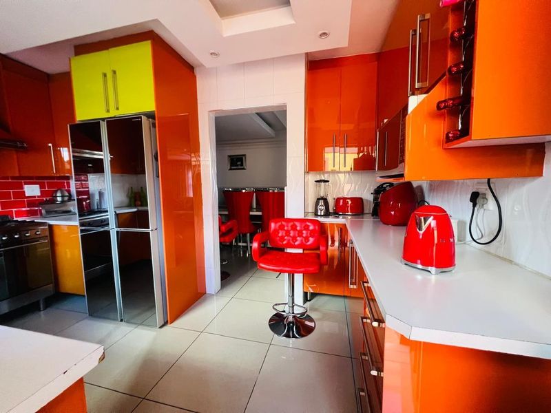 A vibrant kitchen featuring high-gloss orange cabinetry, white countertops, tiled backsplash, and recessed ceiling lighting, with an open doorway leading to the dining area.